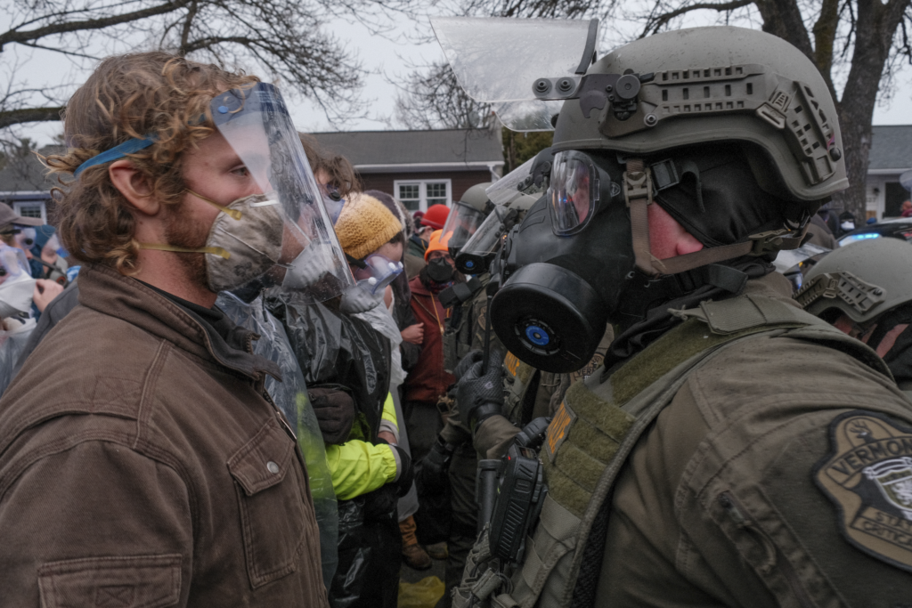 A masked protester face to face with a Vermont State Trooper wearing riot gear and a gas mask on Dorset St. in South Burlington.